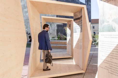 A man carrying a briefcase walks through a walk-in wooden sculpture with text panels in an urban setting