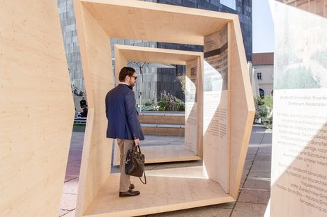 A man carrying a briefcase walks through a walk-in wooden sculpture with text panels in an urban setting