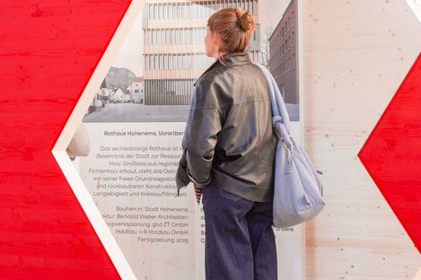 Woman with bun wearing black leather jacket and blue bag standing in front of wall with text and architectural drawing