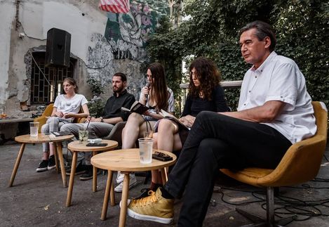 Five people sitting on chairs and stools in an outdoor area with an ivy-covered wall and graffiti in the background, one person speaking into a microphone.