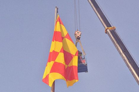 A red and yellow checkered flag flies on a pole next to a crane lifting a work platform with a person into the sky.