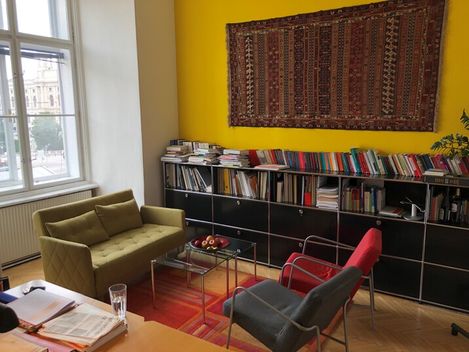 Wohnzimmer mit gelber Wand, Teppich an der Wand, Bücherregal, grünem Sofa, rotem und grauem Sessel sowie Glastisch