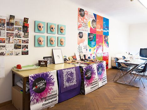 Raum mit Tischen, an denen Poster, Flyer und sechs Masken mit großen Augen hängen, im Hintergrund Stühle und ein alter Fernseher