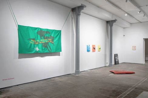 Exhibition room with a green flag on the wall showing a black bird and yellow text, next to three small colorful pictures and a red cushion on the floor.