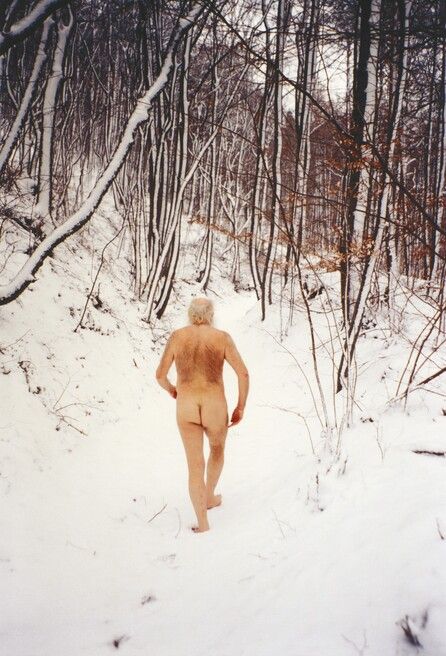 Naked person seen from behind walking barefoot on a snow-covered path through a forest with bare trees