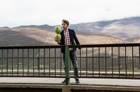 A man stands behind a railing balancing two watermelons stacked in front of a mountainous landscape.