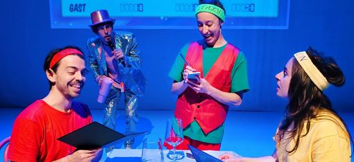A scene from a TV show: The presenter is wearing a glittering silver suit and holding a microphone. The contestants’ scores are displayed in the background. In the foreground, a cheerful waitress is taking the order from two guests.