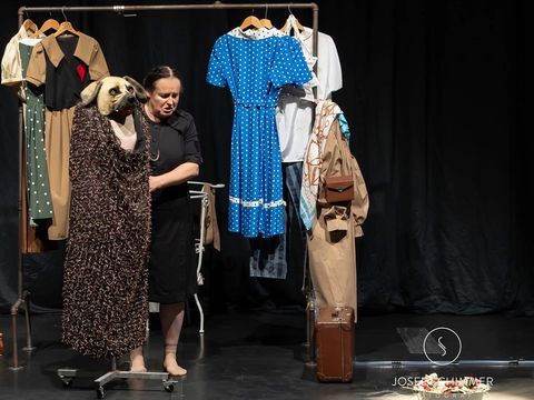 Woman in black dress stands on stage next to clothing rack with various garments and a costume featuring a large dog head.