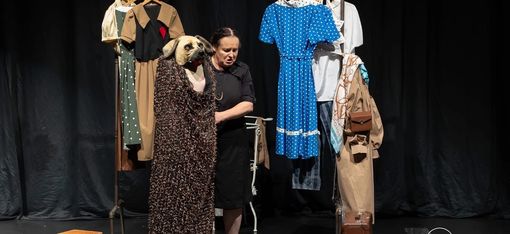 Woman in black dress stands on stage next to clothing rack with various garments and a costume featuring a large dog head.