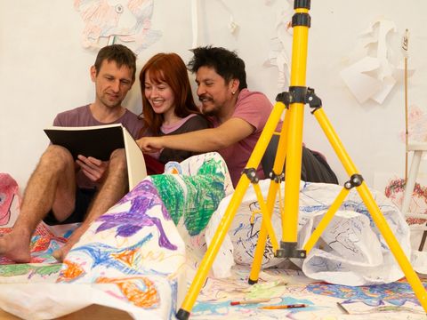 Three people are sitting on the floor, reading from a large book. They are surrounded by a painted canvas.