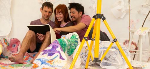 Three people are sitting on the floor, reading from a large book. They are surrounded by a painted canvas.