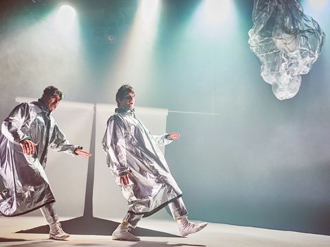 Two men, dressed in quirky silver outfits with round glasses perched on their heads, move across the stage with unusual strides.