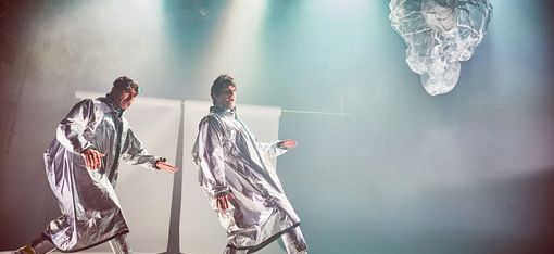 Two men, dressed in quirky silver outfits with round glasses perched on their heads, move across the stage with unusual strides.