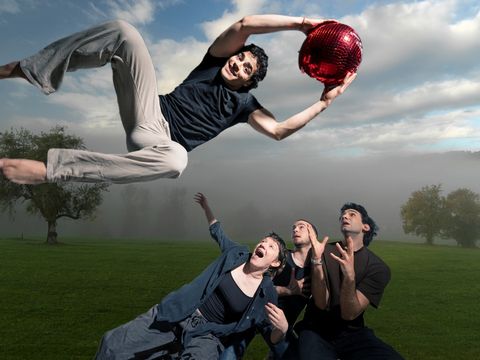 A person, holding a bright red ball, leaps over three people who are looking up at him in admiration.
