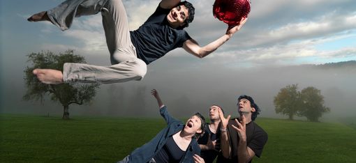 A person, holding a bright red ball, leaps over three people who are looking up at him in admiration.