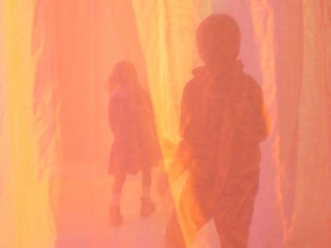 Two people are walking, with their backs to the camera, behind a yellow and pink curtain.