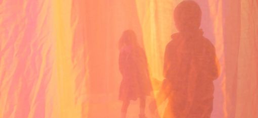 Two people are walking, with their backs to the camera, behind a yellow and pink curtain.