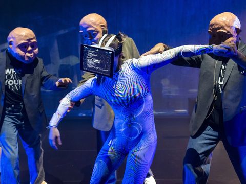 In the centre is a dancer with a laptop held up to her face. Grouped around her are three men wearing masks. An office is projected in the background.
