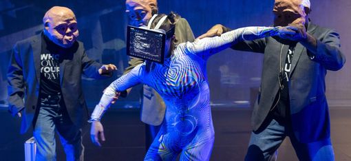 In the centre is a dancer with a laptop held up to her face. Grouped around her are three men wearing masks. An office is projected in the background.