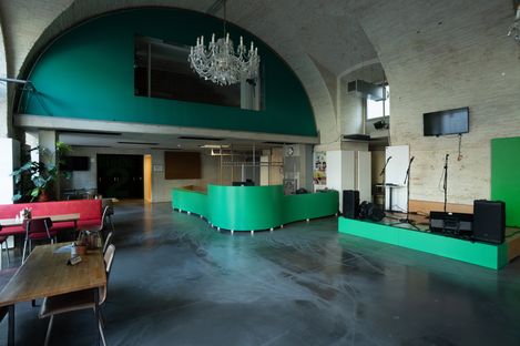 Large function room with a green reception area, a stage, microphones and a chandelier, as well as coffee tables