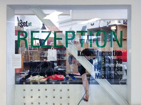 Glaswand mit Aufschrift "REZEPTION" und weiteren Informationen vor einem Empfangstresen mit verschiedenen Gegenständen und einer Person dahinter.