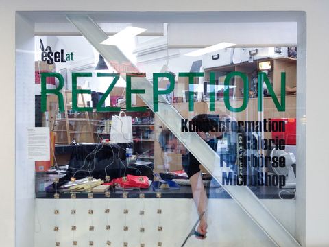 Glaswand mit Aufschrift "REZEPTION" und weiteren Informationen vor einem Empfangstresen mit verschiedenen Gegenständen und einer Person dahinter.