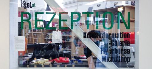 Glaswand mit Aufschrift "REZEPTION" und weiteren Informationen vor einem Empfangstresen mit verschiedenen Gegenständen und einer Person dahinter.