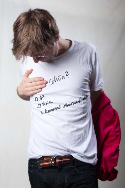 Person mit braunen Haaren schaut auf ein weißes T-Shirt mit handgeschriebenem Text und hält eine rote Jacke in der linken Hand.