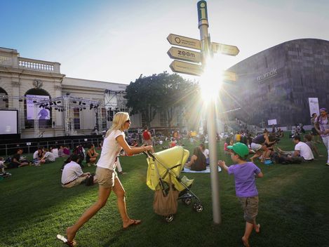 Frau schiebt Kinderwagen auf einer Wiese mit mehreren sitzenden Menschen vor einem modernen und einem historischen Gebäude bei Sonnenlicht