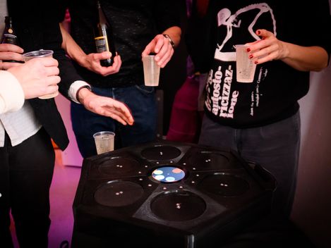 Mehrere Personen halten Getränke und stehen um einen runden schwarzen Tisch mit mehreren Vertiefungen und einem beleuchteten Display in der Mitte.
