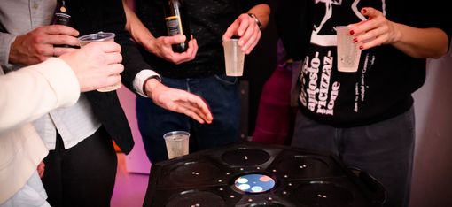 Mehrere Personen halten Getränke und stehen um einen runden schwarzen Tisch mit mehreren Vertiefungen und einem beleuchteten Display in der Mitte.