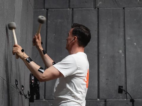 Mann mit weißem T-Shirt spielt mit zwei Schlägeln an einer Wand aus grauen, rechteckigen Platten.