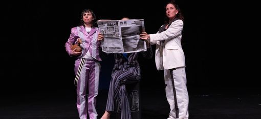 Three performers in colorful suits on stage: one hides behind a newspaper, while the others hold it in place.