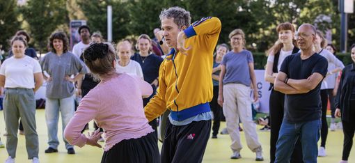 Eine Gruppe von Menschen steht auf einem gelben Boden im Freien, zwei Personen in der Mitte führen eine Tanzbewegung aus, während die anderen zuschauen.