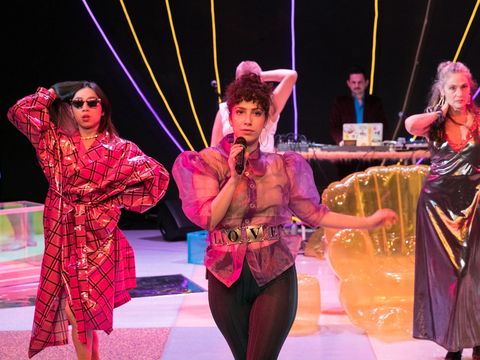 Vier Frauen stehen auf einer Bühne und präsentieren Kostüme, eine der Frauen hält ein Mikrofon in der Hand. Im Hintergrund stehen zwei Männer am Mischpult.