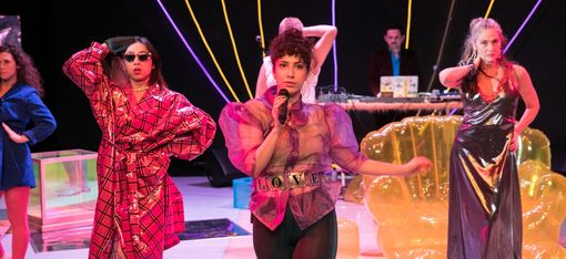 Vier Frauen stehen auf einer Bühne und präsentieren Kostüme, eine der Frauen hält ein Mikrofon in der Hand. Im Hintergrund stehen zwei Männer am Mischpult.