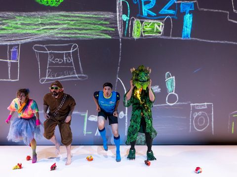 Four figures (two humans, an elf, a dragon) are standing on a stage: They are wearing extravagant costumes, such as a glittering skirt, a full-body plush suit with elf ears, and a sports outfit. The green dragon looks friendly.