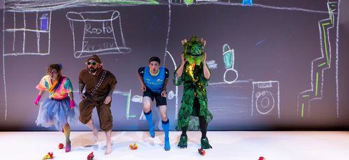 Four figures (two humans, an elf, a dragon) are standing on a stage: They are wearing extravagant costumes, such as a glittering skirt, a full-body plush suit with elf ears, and a sports outfit. The green dragon looks friendly.