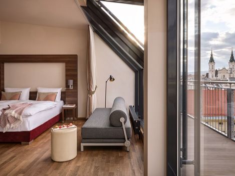 Hotelzimmer mit Doppelbett, grauem Sofa, runden Hocker und großem Fenster mit Blick auf die Dächer und Türme einer Stadt