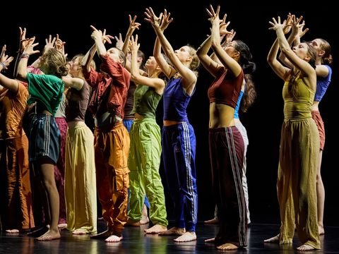 A group of young people are standing in a choreographed pose: they are stretching their arms upwards and spreading their fingers. Their gaze is directed towards their hands.