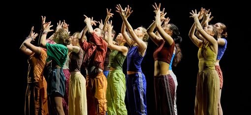 A group of young people are standing in a choreographed pose: they are stretching their arms upwards and spreading their fingers. Their gaze is directed towards their hands.