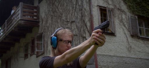 Frau mit Schutzbrille und Gehörschutz hält eine Pistole mit ausgestreckten Armen vor einem Haus mit Balkon und Fenster