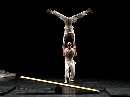 Two people are performing an acrobatic routine to perfection: one person is doing a handstand on the other’s arms and is in the process of spreading their legs into the splits.