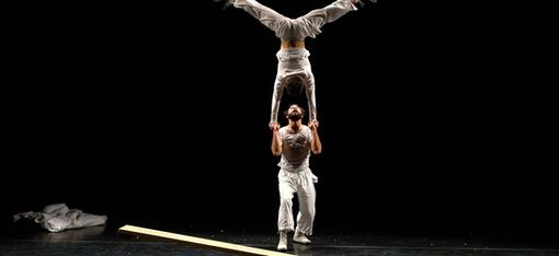 Two people are performing an acrobatic routine to perfection: one person is doing a handstand on the other’s arms and is in the process of spreading their legs into the splits.