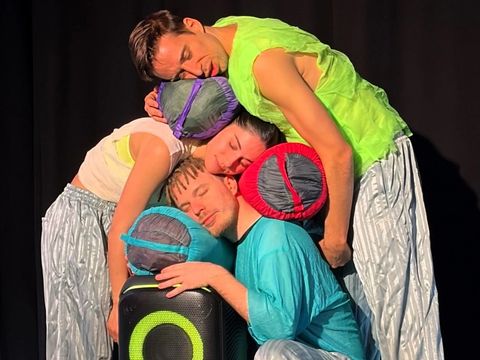 Three dancers have closed their eyes and stacked their heads and sleeping bags on top of each other.
