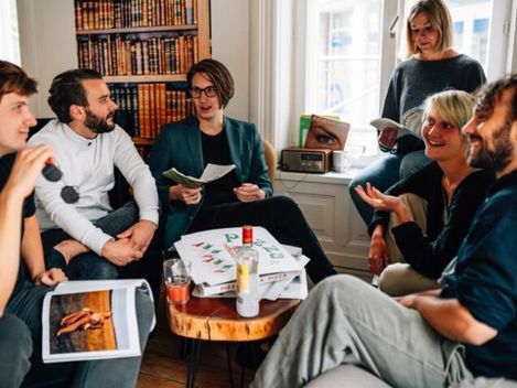 Fünf Personen sitzen in einem Wohnzimmer um einen kleinen Holztisch mit Pizzakartons und Getränken und unterhalten sich.