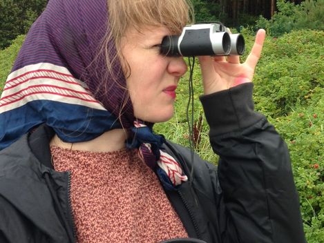 Person wearing a headscarf holding binoculars to their eyes and a cup in the other hand against a background of greenery.