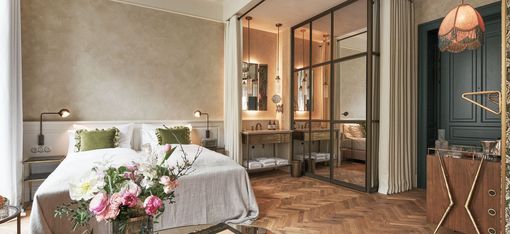 Ein gemütliches Schlafzimmer verfügt über ein weiches Doppelbett mit grünen Kissen und einer grauen Decke. Frische Blumen auf einem Glastisch und eine warme Beleuchtung schaffen eine ruhige Atmosphäre.