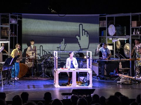 Six musicians stand in a semicircle on stage with their instruments. In the centre of the stage, a woman in a white lab coat is sitting at a laboratory table. The set consists of metal shelves and resembles a garage filled with useful and forgotten items. A large screen hangs in the background.