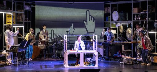 Six musicians stand in a semicircle on stage with their instruments. In the centre of the stage, a woman in a white lab coat is sitting at a laboratory table. The set consists of metal shelves and resembles a garage filled with useful and forgotten items. A large screen hangs in the background.